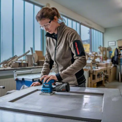 Mulher lixa uma porta branca numa oficina com lixadora azul e preta