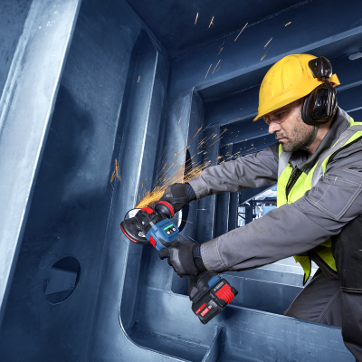 Trabalhador com capacete amarelo e colete refletor a usar rebarbadora em estrutura metálica azul