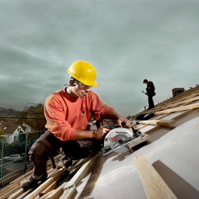 Dois trabalhadores num telhado, um corta madeira com serra circular Makita e usa capacete amarelo.