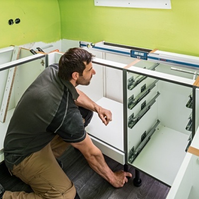 Homem a instalar móveis de cozinha brancos com puxadores metálicos num espaço com paredes verdes e chão escuro