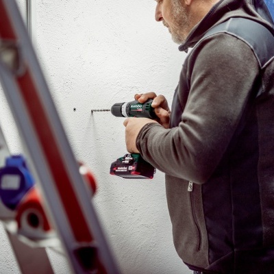 Homem a usar furadeira sem fios verde e preta numa parede branca.