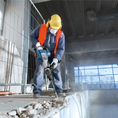 Trabalhador de construção com capacete e colete laranja usando martelo pneumático em obra interior