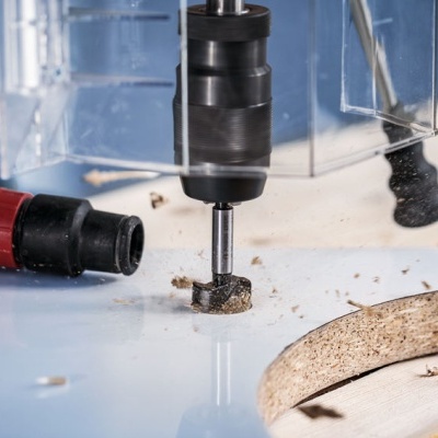 Máquina de perfuração a trabalhar em madeira com aparas e tubos ao fundo