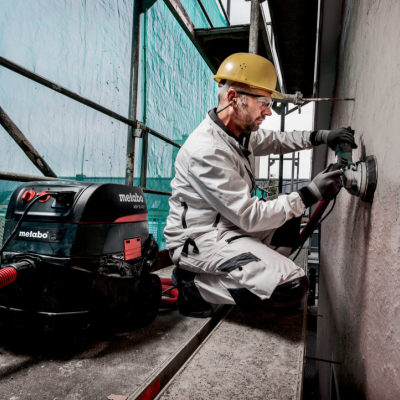 Homem a lixar parede com ferramenta elétrica num estaleiro de construção com máquina Metabo de aspiração.