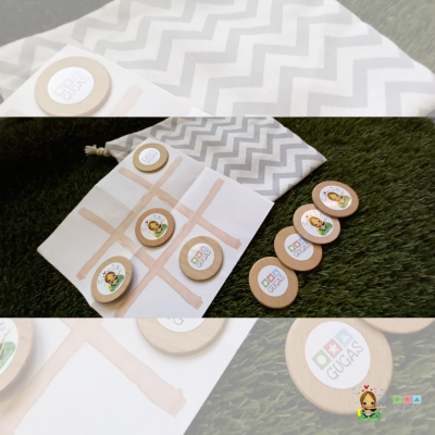 Jogo da velha em madeira com peças etiquetadas GUGAS sobre relva verde e pano com padrão ziguezague cinza e branco