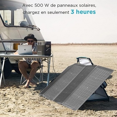 Painéis solares dobráveis pretos na areia perto de um veículo recreativo e uma pessoa a trabalhar num portátil numa mesa portátil