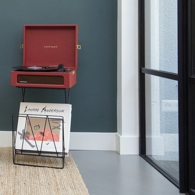 Aparelho de vinil vermelho escuro LONON com caixa para discos em suporte preto num chão cinza ao lado de parede verde