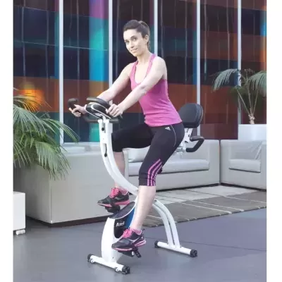 Mulher a usar bicicleta estática branca num espaço interior com sofás brancos e plantas.