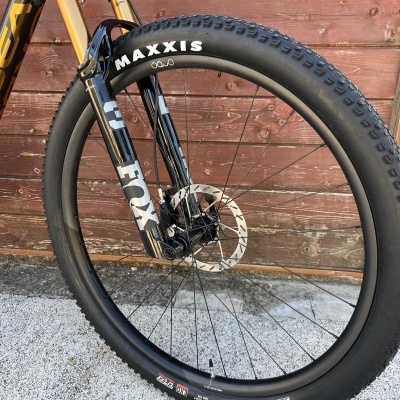 Roda frontal de bicicleta com pneu Maxxis e garfo FOX encostada a parede de madeira vermelha