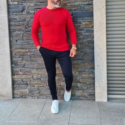 Homem com camisola vermelha, calças pretas e sapatilhas brancas junto a parede de pedra.