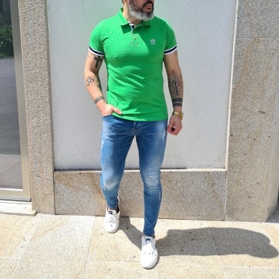 Homem vestido com polo verde, jeans azuis e sapatilhas brancas em fundo de parede branca com moldura de pedra