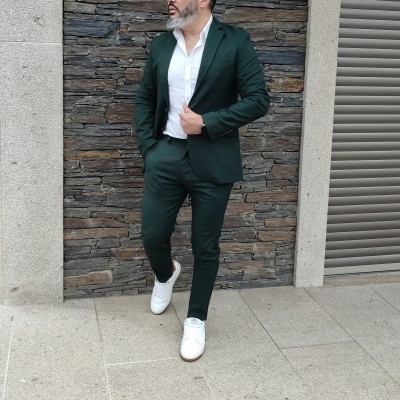Homem com fato verde escuro e camisa branca junto a parede de pedra.