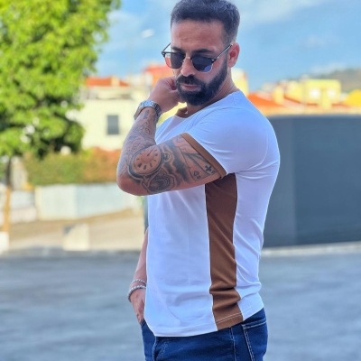 Homem com camiseta branca e castanha, jeans azul, usando óculos de sol em ambiente externo