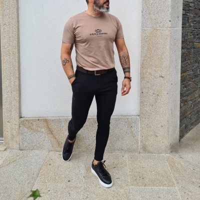 Homem com t-shirt bege, calças pretas e sapatilhas pretas em ambiente exterior de pedra