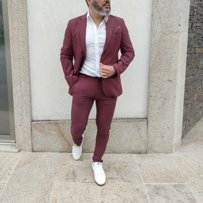 Homem com fato bordô, camisa branca e sapatos brancos junto a parede de pedra clara