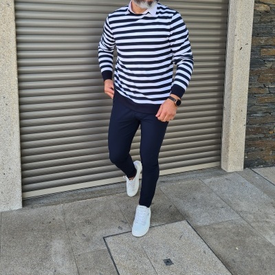 Homem com camisa riscada azul e branca, calças azuis escuras e sapatilhas brancas junto a porta metálica.