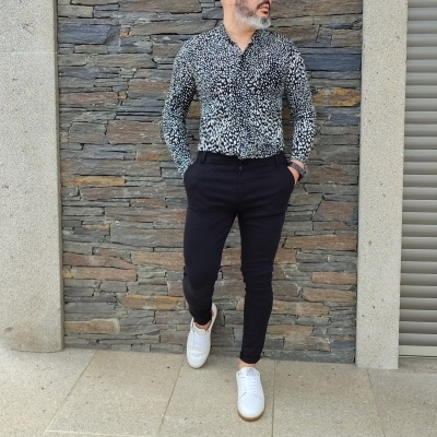 Homem com camisa estampada preto e branco, calças pretas e sapatilhas brancas junto à parede de pedra cinzenta