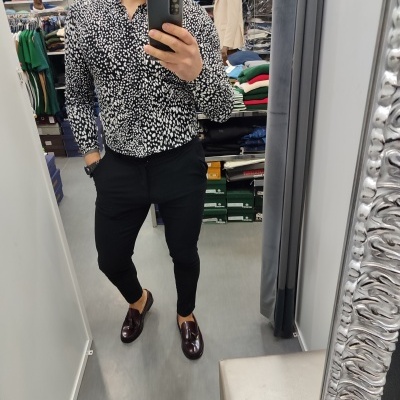 Homem a tirar selfie no espelho de loja, camisa preta com padrão branco, calças pretas, sapatos castanho-escuros