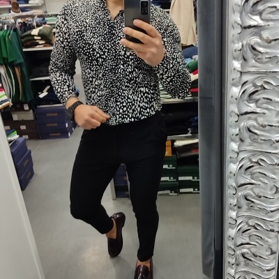 Homem numa loja de roupa com camisa preta padrão branco, calças pretas e sapatos castanhos escuros