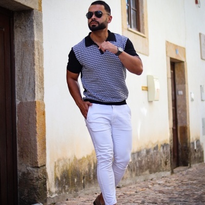 Homem com camisa preta e branca, calças brancas e sapatos bege em rua de pedra