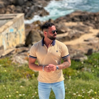 Homem de camisa polo bege e calças de ganga junto ao mar