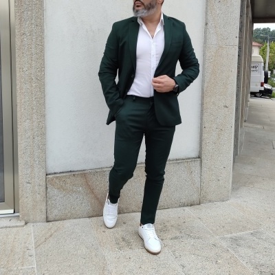 Homem com fato verde-escuro, camisa branca e sapatos brancos junto a parede clara