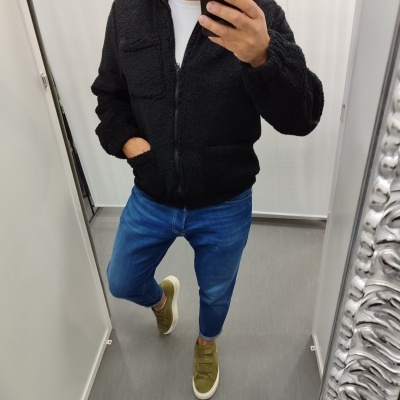 Homem a vestir casaco preto felpudo, calças jeans azuis e sapatilhas verdes em frente a um espelho prateado.