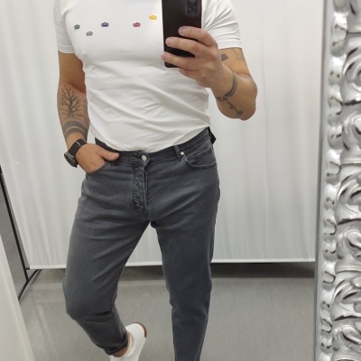 Homem com t-shirt branca, jeans cinza escuro e sapatilhas brancas a tirar selfie num espelho