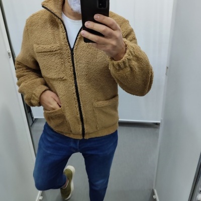 Casaco castanho sherpa com fecho e bolsos, calças azuis e sapatos verdes num provador branco.