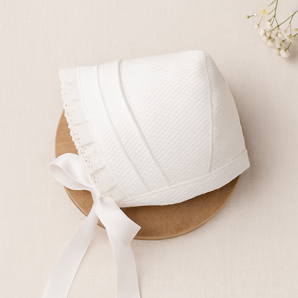Chapéu branco de bebé com fita de cetim e renda, em fundo claro com prato de madeira.