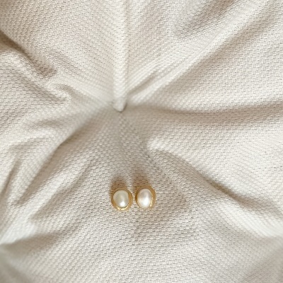 Brincos de pérola oval e metal dourado sobre tecido branco texturizado