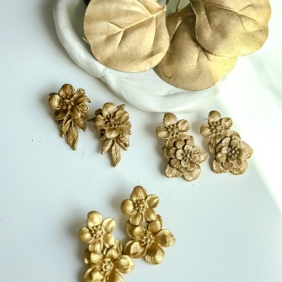 Três pares de brincos florais dourados sobre superfície branca com folhas douradas decorativas