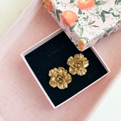 Brincos dourados em forma de flor numa caixa de veludo preta dentro de uma caixa rosa decorada com laranjas e flores.