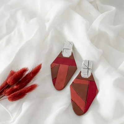 Brincos geométricos em tons vermelhos e terra sobre pano branco com ramo de plantas secas vermelhas