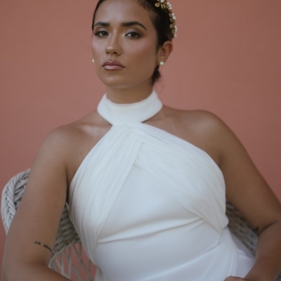 Mulher com vestido branco de gola alta e detalhe cruzado, sentada numa cadeira de vime, fundo rosa claro