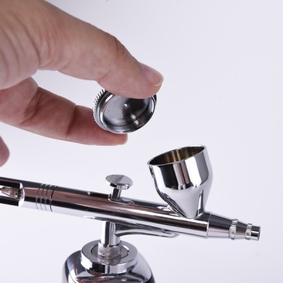 Pistola de aerógrafo metálica prateada com copo para tinta e tampa rosqueada.