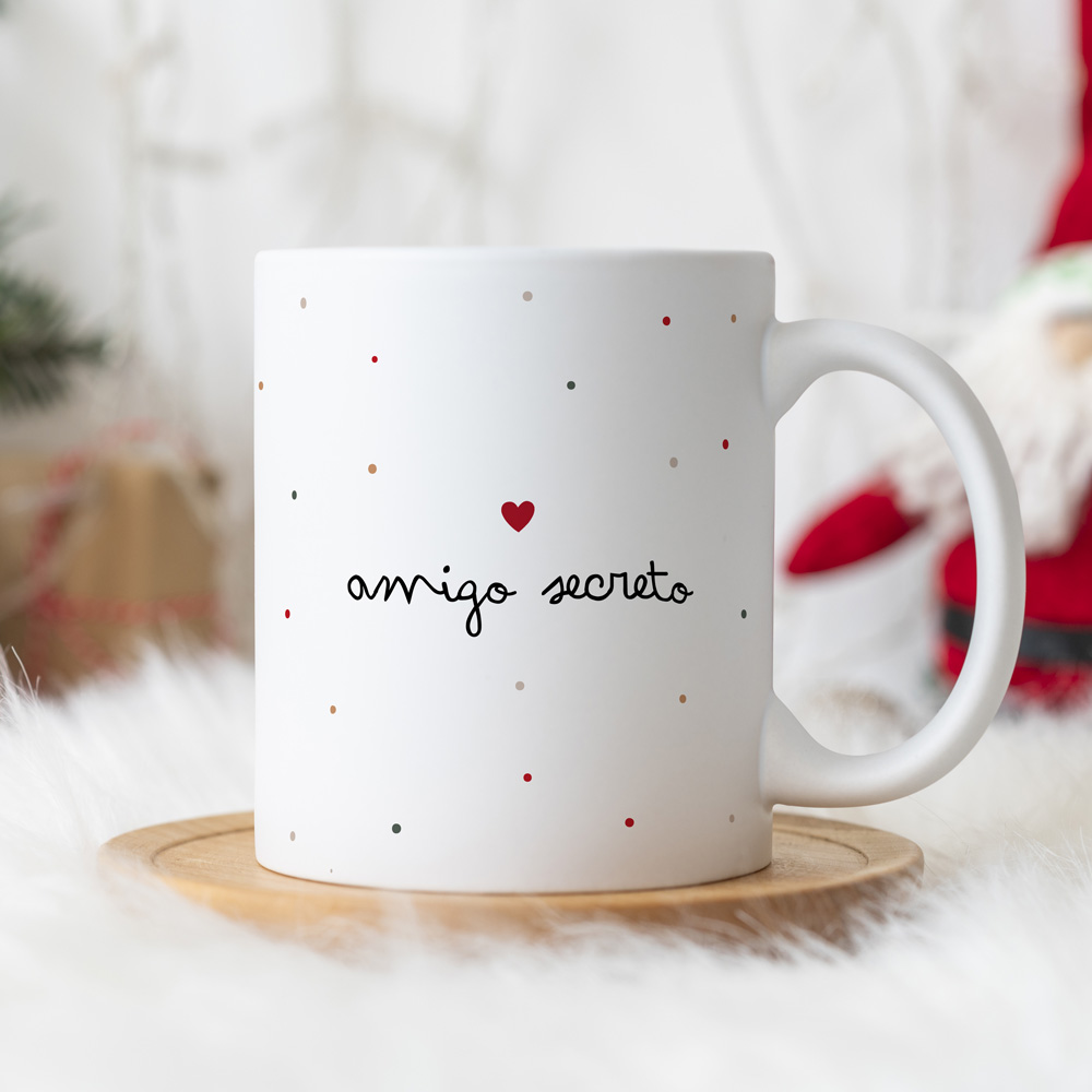 Caneca branca com texto 'amigo secreto' e padrão de pontos coloridos, sobre base de madeira, cenário natalício desfocado