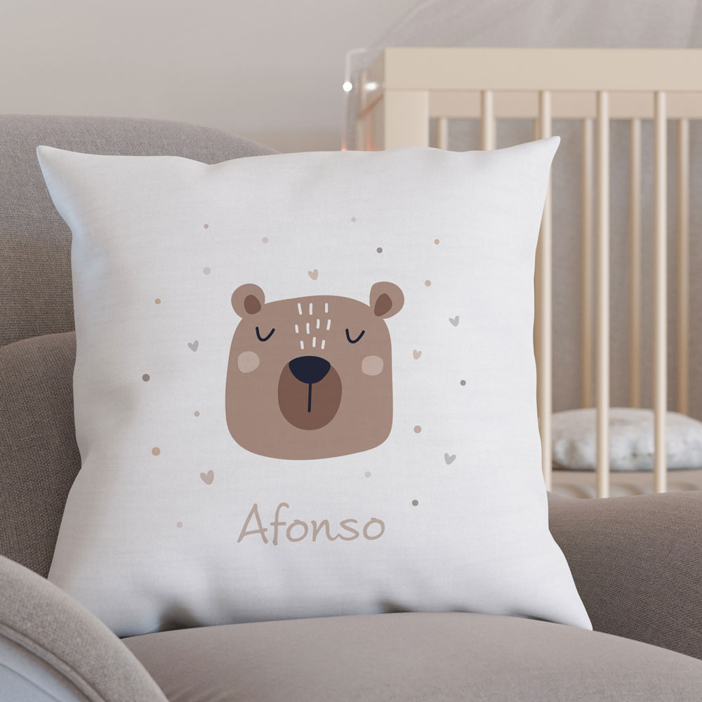 Almofada branca com rosto de urso e nome Afonso num sofá junto a um berço branco