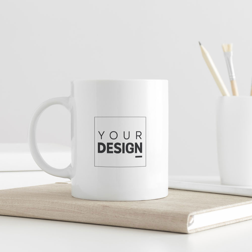 Caneca branca de cerâmica com design YOUR DESIGN, sobre livro bege em mesa branca com lápis e pincéis