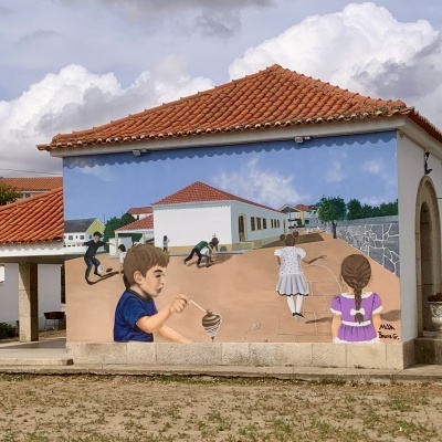 Mural colorido com crianças a brincar pintado numa parede exterior de edifício branco com telhado de telhas vermelhas