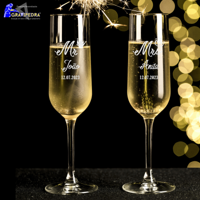 Duas flautas de champanhe gravadas com textos e datas em fundo preto com luzes desfocadas douradas.