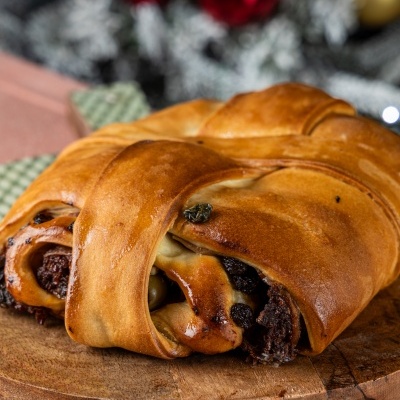 Dois pães doces entrelaçados com recheio de chocolate e passas numa tábua de madeira com decoração natalícia ao fundo.