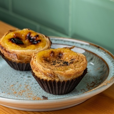 Dois pastéis de nata em prato azul sobre mesa de madeira clara com fundo de azulejos verdes