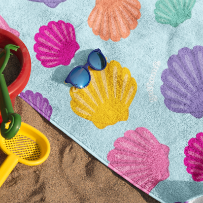 Toalha de praia azul clara com conchas coloridas, óculos de sol, balde e pá na areia