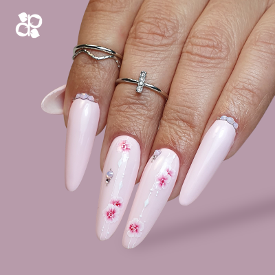 Unhas rosa com decoração floral e anéis prateados sobre fundo lilás