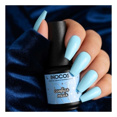 Unhas pintadas de azul claro segurando verniz INOCOS sobre tecido azul