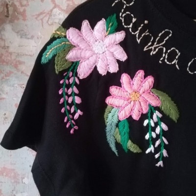 T-shirt preta com flores bordadas rosas e folhas verdes no ombro, texto branco bordado