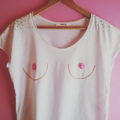 T-shirt branca com estampado de contorno de seios e pérolas nas mangas, pendurada num cabide de madeira frente a fundo cor-de-rosa.