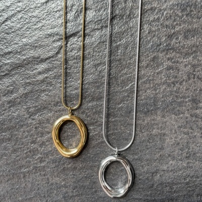 Dois colares com pingentes em forma de anel oval irregular, um dourado e outro prateado, sobre fundo cinza escuro texturado.