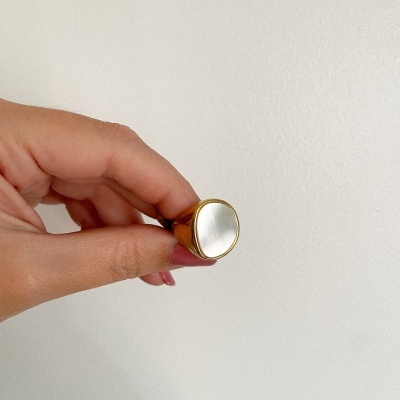 Anel dourado com placa oval branca na parte superior, mão segurando, fundo branco.